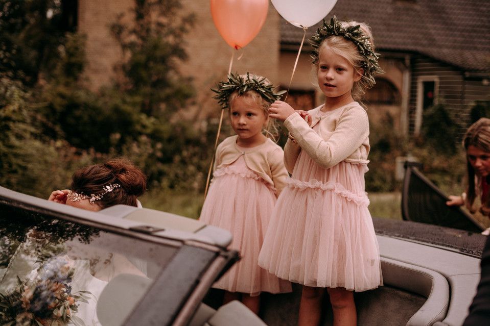 twee kleine meisjes in roze jurken en bloemenkransen staan achterin een auto