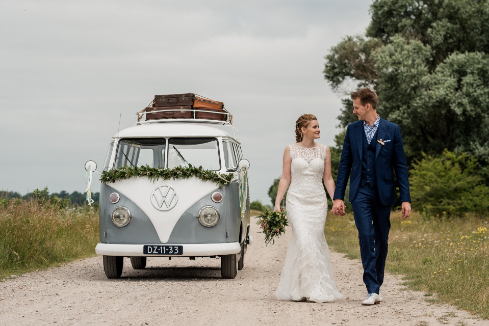 een bruid en bruidegom lopen op een zandweg bij een vw camperbusje