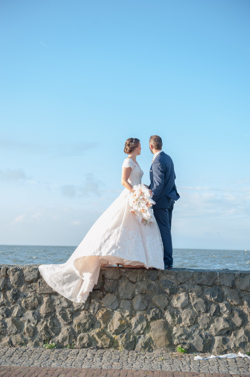 een bruid en bruidegom staan op de rand van een muur bij de oceaan