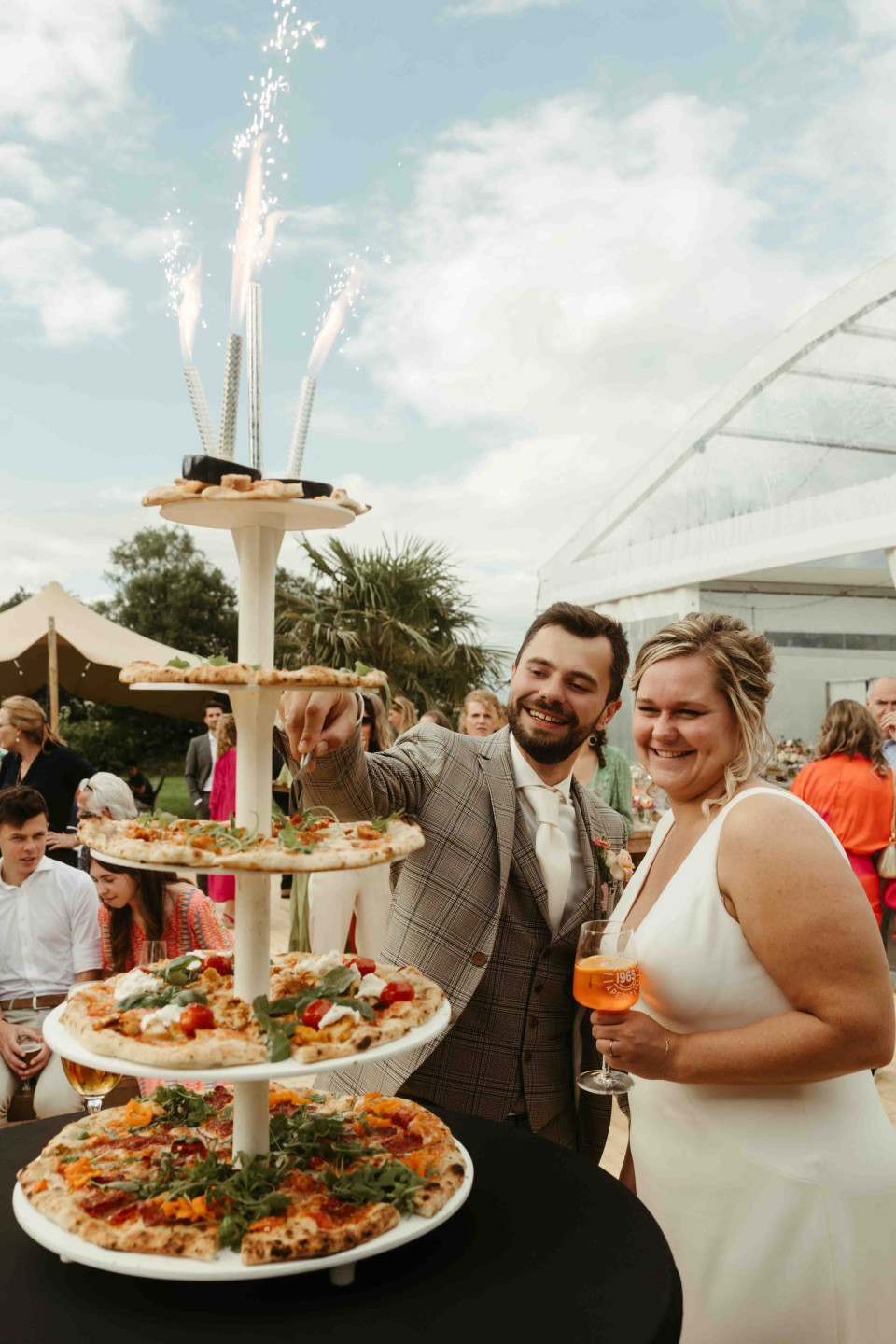 een man en een vrouw die voor een drie-laags schaal met pizza's staan en vuurwerk in de lucht hebben