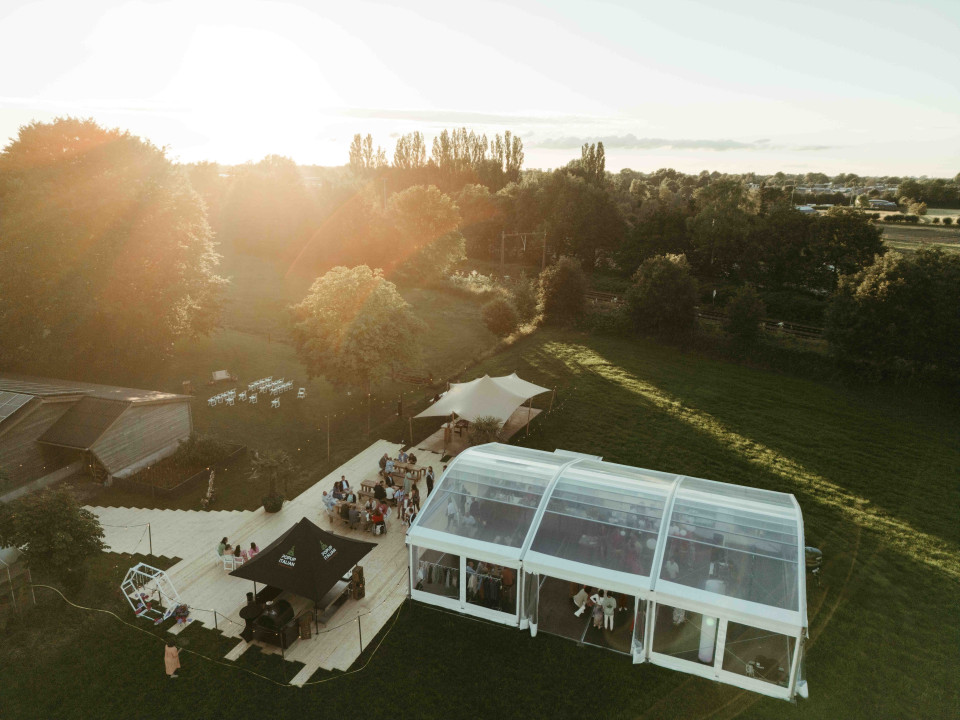een grote groep mensen die buiten staan van een grote witte tent in het midden van een grasveld met bomen op de achtergrond