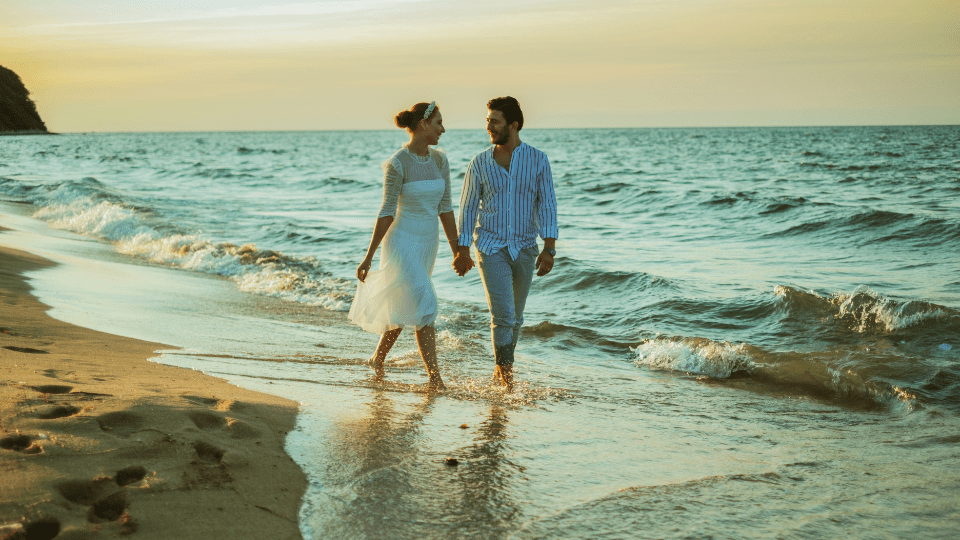 een man en een vrouw die hand in hand over het strand lopen en weg van het water lopen met de zon die ondergaat op de achtergrond