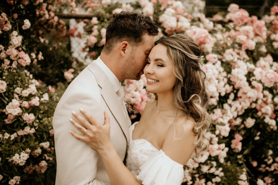 een bruid en bruidegom die voor een struik met roze bloemen staan
