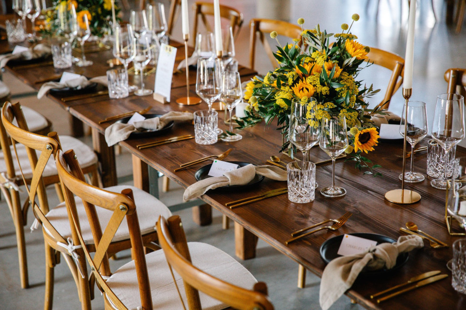 een lange houten tafel met een stel glazen en bloemen erop met plaatskaarten op elke stoel
