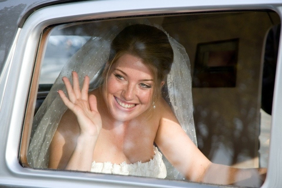 een lachende bruid zwaait vanaf de achterbank van een auto