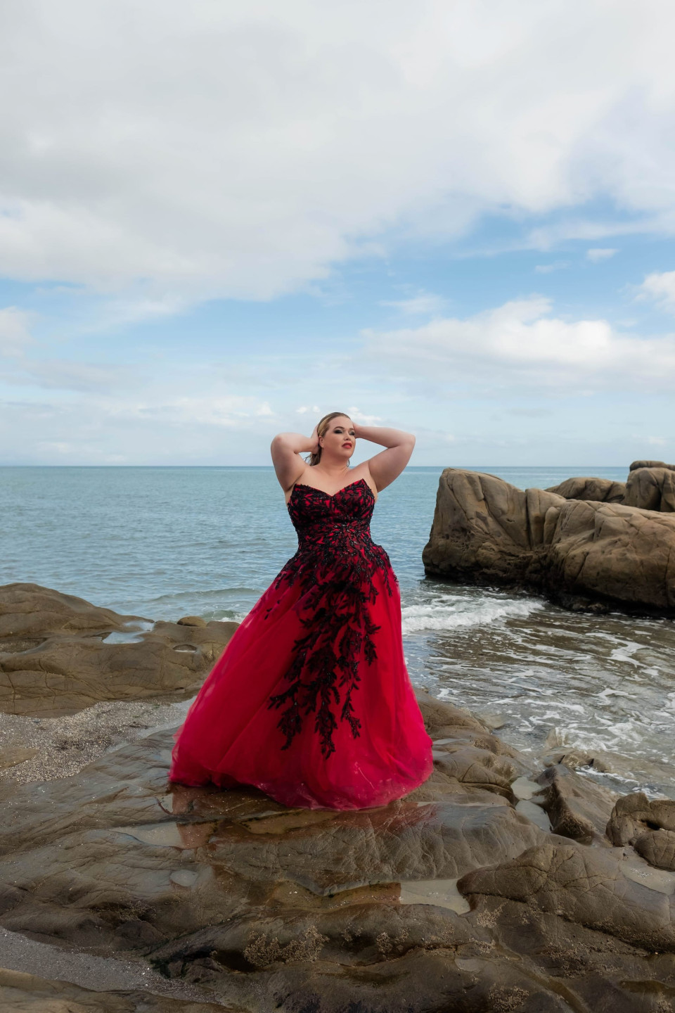 een vrouw in een rode jurk die op rotsen bij de oceaan staat