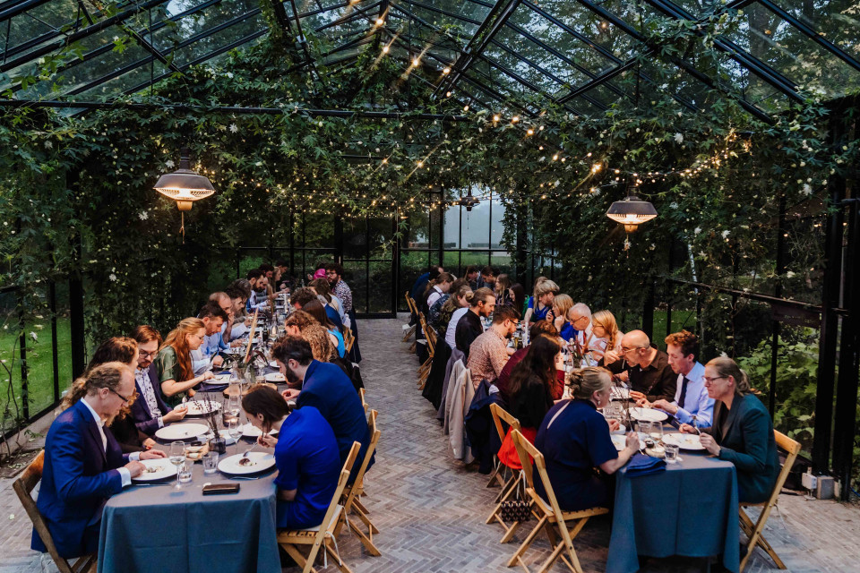 een grote groep mensen die aan tafels zitten in een kas, eten en wijn drinken en wijnglazen op de tafel