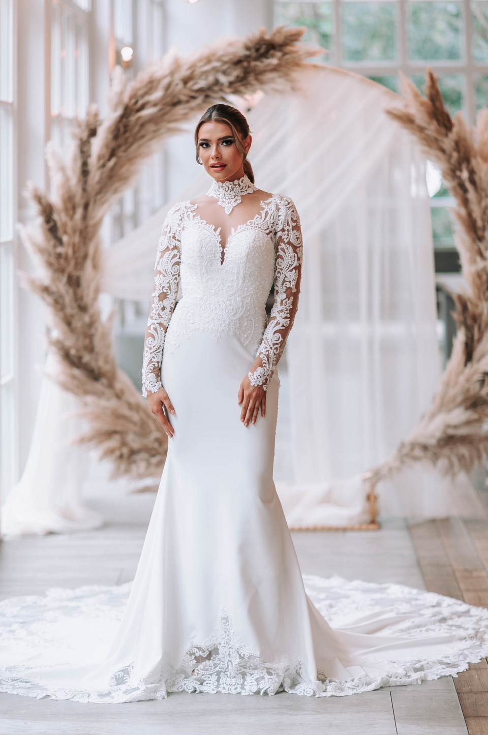 een bruid in een lange mouwen trouwjurk poseert voor een grote krans