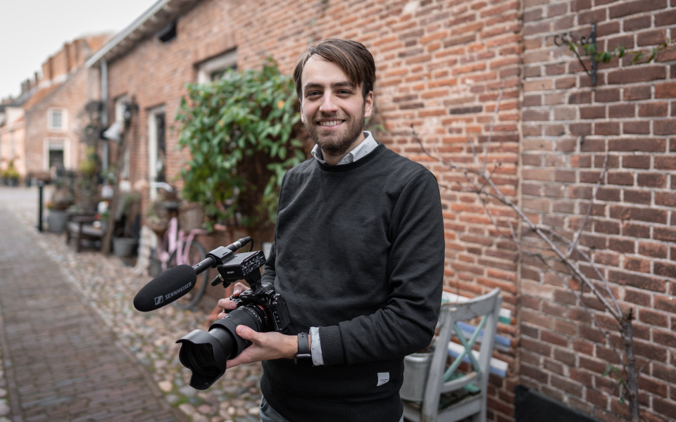 een man die een camera vasthoudt en lacht voor een bakstenen gebouw
