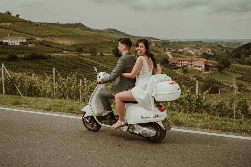een man en een vrouw die op een scooter rijden op een weg met een wijngaard op de achtergrond en heuvels in de verte
