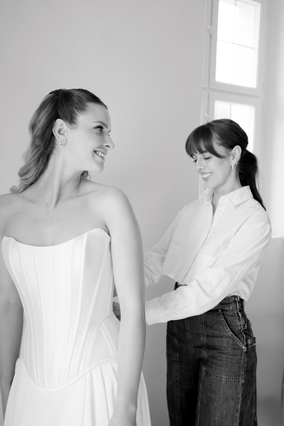een vrouw die een vrouw helpt haar trouwjurk aan te trekken in een zwart-witfoto met een raam op de achtergrond
