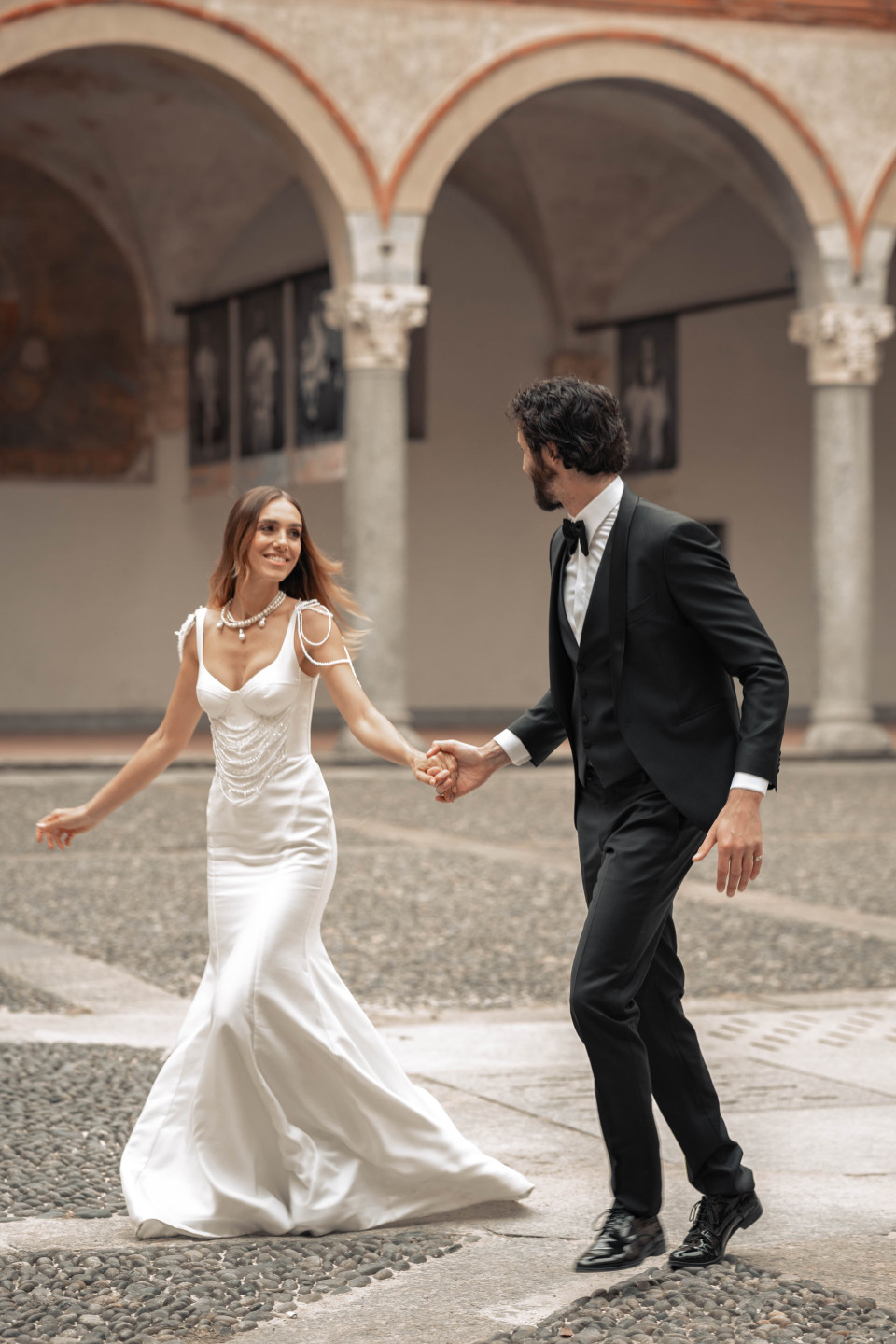 een man en een vrouw in een trouwjurk die hand in hand lopen door een binnenplaats met bogen op de achtergrond